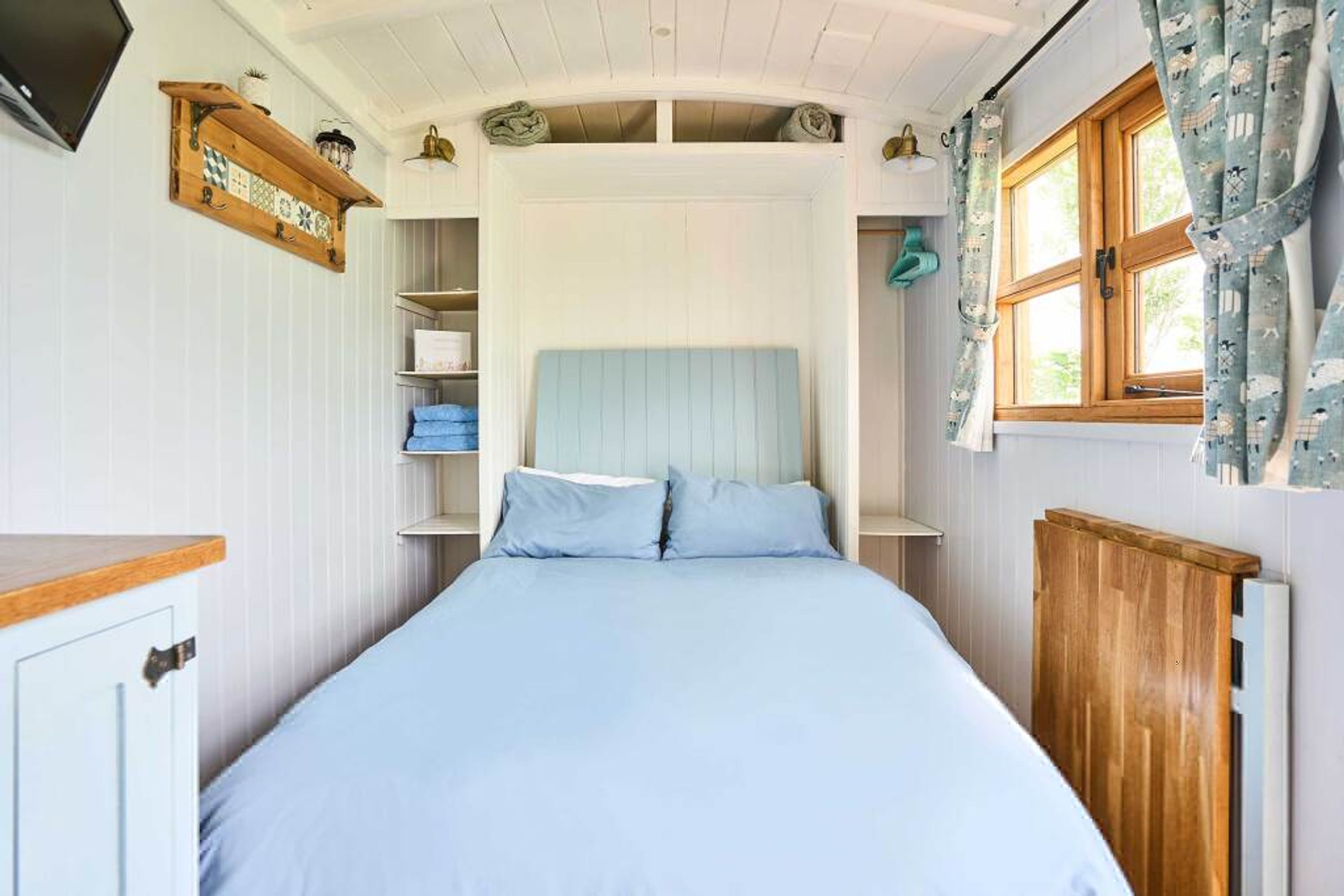 A bedroom with a bed, wooden headboard, a shelf with towels, a window with curtains, and a TV mounted on the wall.
