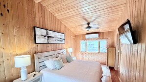 Ein Schlafzimmer mit Holzwandverkleidung, einem Bett, Nachttischlampen, einem Deckenventilator und einem an der Wand befestigten Fernseher.
