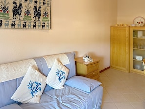 A room with a light blue sofa, patterned cushions, a wooden cabinet with glass doors, and a wall-mounted tapestry. - Coniglio Beach by Interhome (Villasimius)