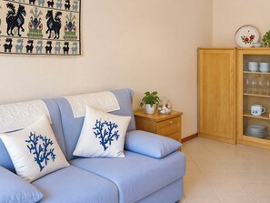 A living room with a sofa, pillows, a small table with a plant, a cabinet with dishes, and a wall with a decorative print. - Coniglio Beach by Interhome (Villasimius)