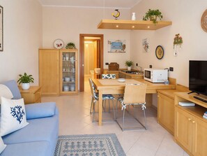 A modern living room with a sofa, dining table, chairs, a TV, and a kitchen area with a microwave and sink. - Coniglio Beach by Interhome (Villasimius)