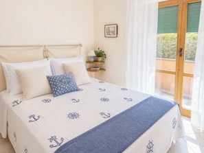 A bed with nautical-themed bedding, a bedside table with a lamp and a plant, and a window with a view of greenery. - Coniglio Beach by Interhome (Villasimius)