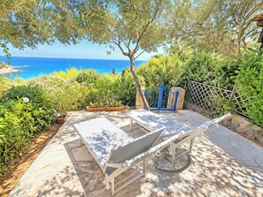 An outdoor seating area with two lounge chairs and a table, surrounded by greenery and overlooking a body of water. - Porto Luna 1 by Interhome (Villasimius)
