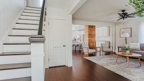 Una scala conduce a un bagno di servizio, una colonna in legno, una zona pranzo con sedie, un divano e una ventola a soffitto.