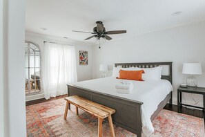 Une chambre à coucher comprenant un lit, un banc, un ventilateur de plafond, une table de chevet et une fenêtre avec des rideaux.