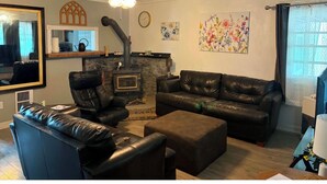 A living room with a black leather sofa, a black leather chair, a ceiling fan, a fireplace, and a large window. - Fantastic 4-bedroom house in quaint Alturas for your getaway Centrally located!! (Alturas)