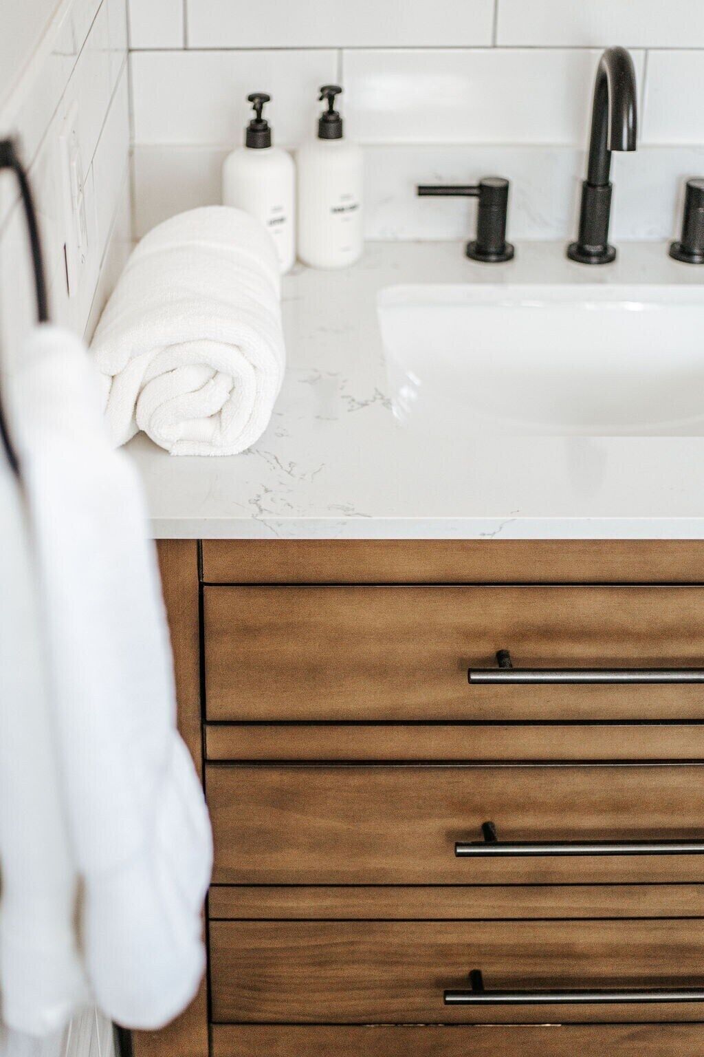 A bathroom with a white sink, black faucet, and two soap dispensers. There are three wooden drawers with black handles.