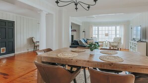 Un vaste espace dédié à la salle à manger, avec une grande table en bois, de nombreuses chaises, un lustre et une vue sur un salon comprenant un canapé et des fauteuils.