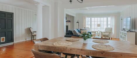 A spacious dining area with a large wooden table, multiple chairs, a chandelier, and a view of a living room with a sofa and chairs.