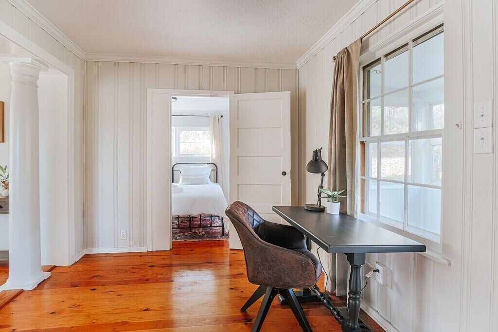 Una habitación con piso de madera, un escritorio con lámpara, una silla y una cama con cabecera blanca.