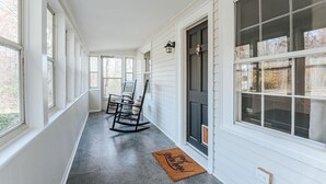 Une véranda couverte aux lambris blancs, une porte noire et un fauteuil à bascule.