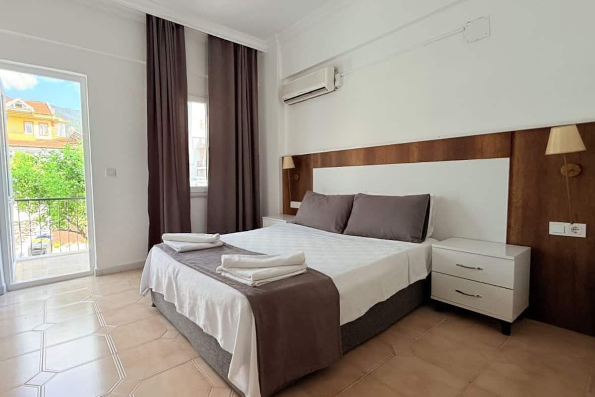Habitación de hotel con cama, mesita de noche, ventana con cortinas y vista a edificios.