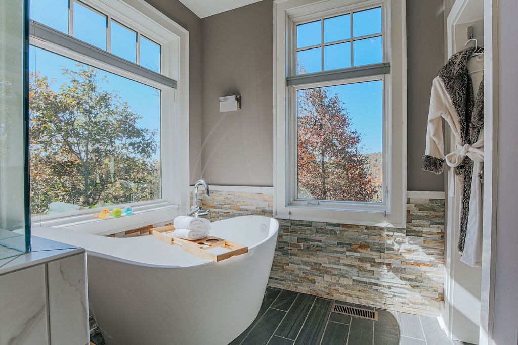 A modern bathroom with a freestanding bathtub, large windows, and a stone backsplash.