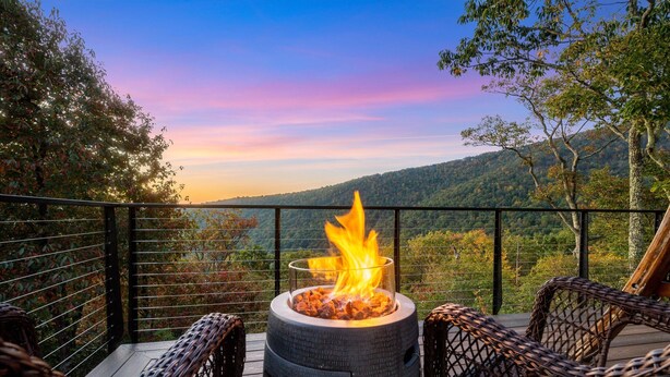A balcony with wicker chairs, a fire pit, and a scenic view of a forested mountain range. - Art & Soul Main House (Nellysford)