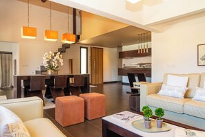 Ein modernes Wohnzimmer mit einer Couch, Hocker, einem Couchtisch und einem Essbereich, wobei sich im Hintergrund eine Küche befindet.