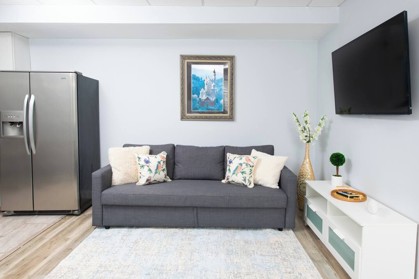Un salon moderne avec un canapé gris, un tableau encadré, un téléviseur à écran plat, un réfrigérateur et une petite plante.