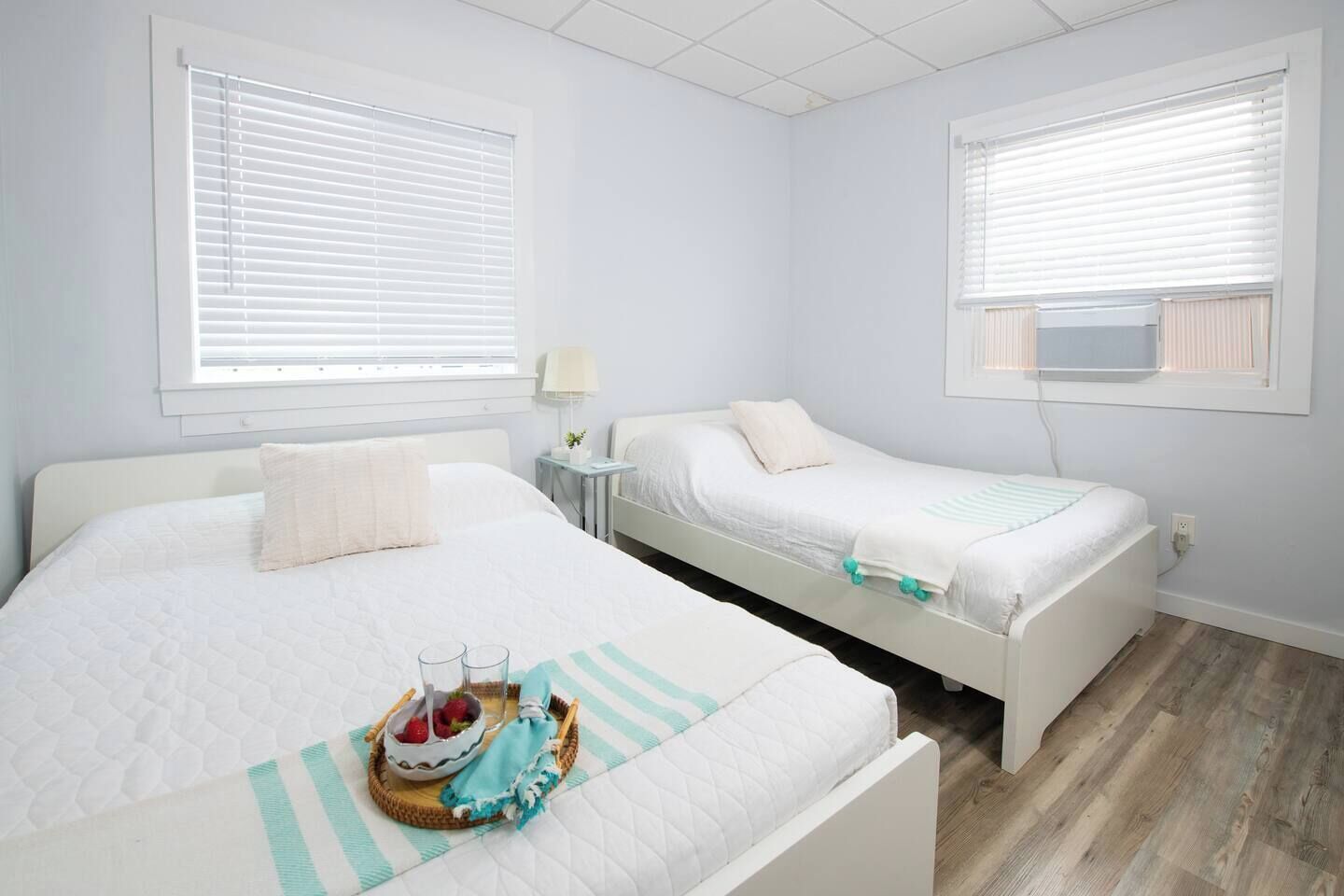A hotel room with two beds, a bedside table with a lamp, and a small table with a fruit bowl and glasses.