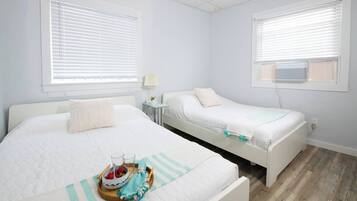 A hotel room with two beds, a bedside table with a lamp, and a small table with a fruit bowl and glasses.