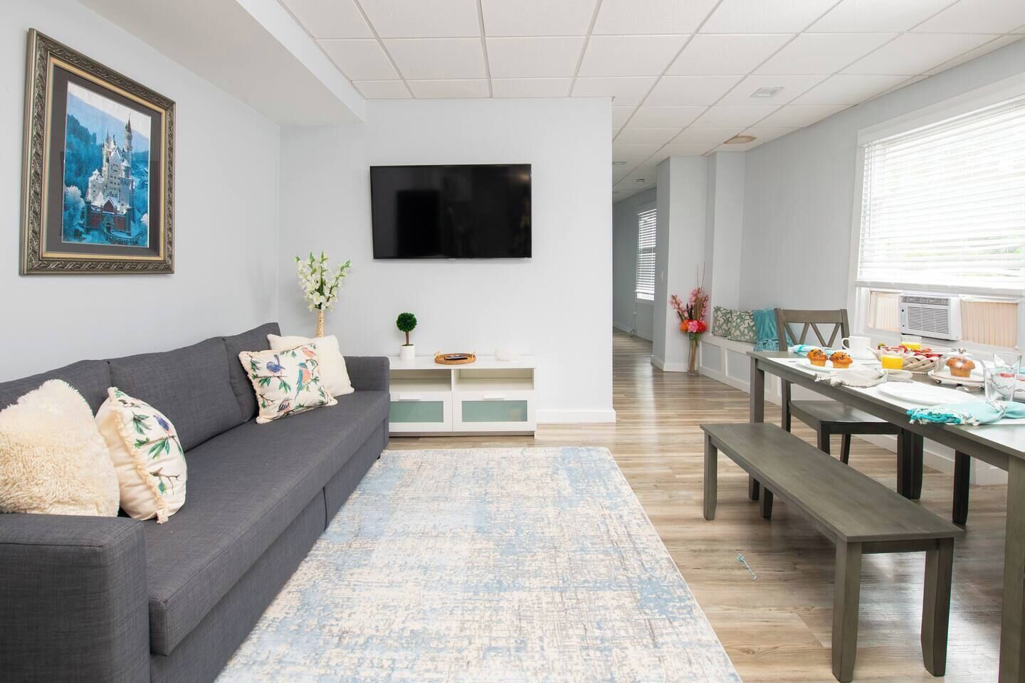 Un salon avec un canapé gris, un téléviseur à écran plat, une table à manger avec des aliments pour le petit-déjeuner et un tableau encadré au mur.