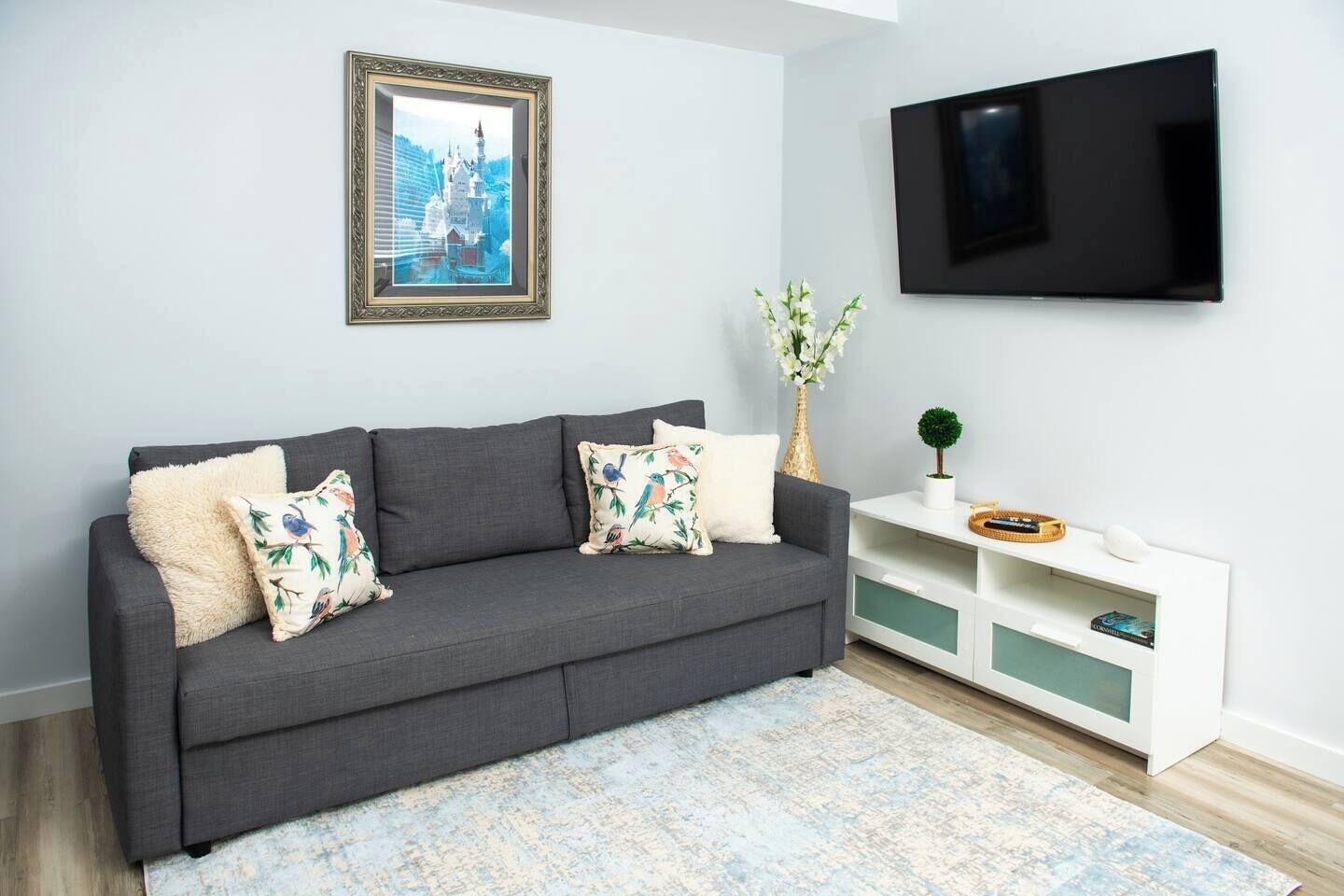 A modern living room with a grey sofa, floral cushions, a framed picture, a TV, and a white console with decorative items.