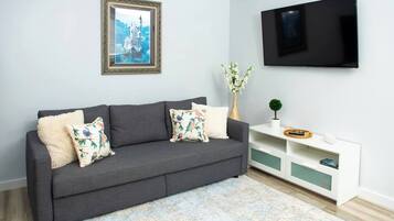 A modern living room with a grey sofa, floral cushions, a framed picture, a TV, and a white console with decorative items.