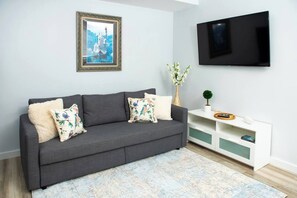 Un salon moderne avec un canapé gris, des coussins à motifs floraux, un tableau encadré, un téléviseur et une console blanche agrémentée d’objets décoratifs.