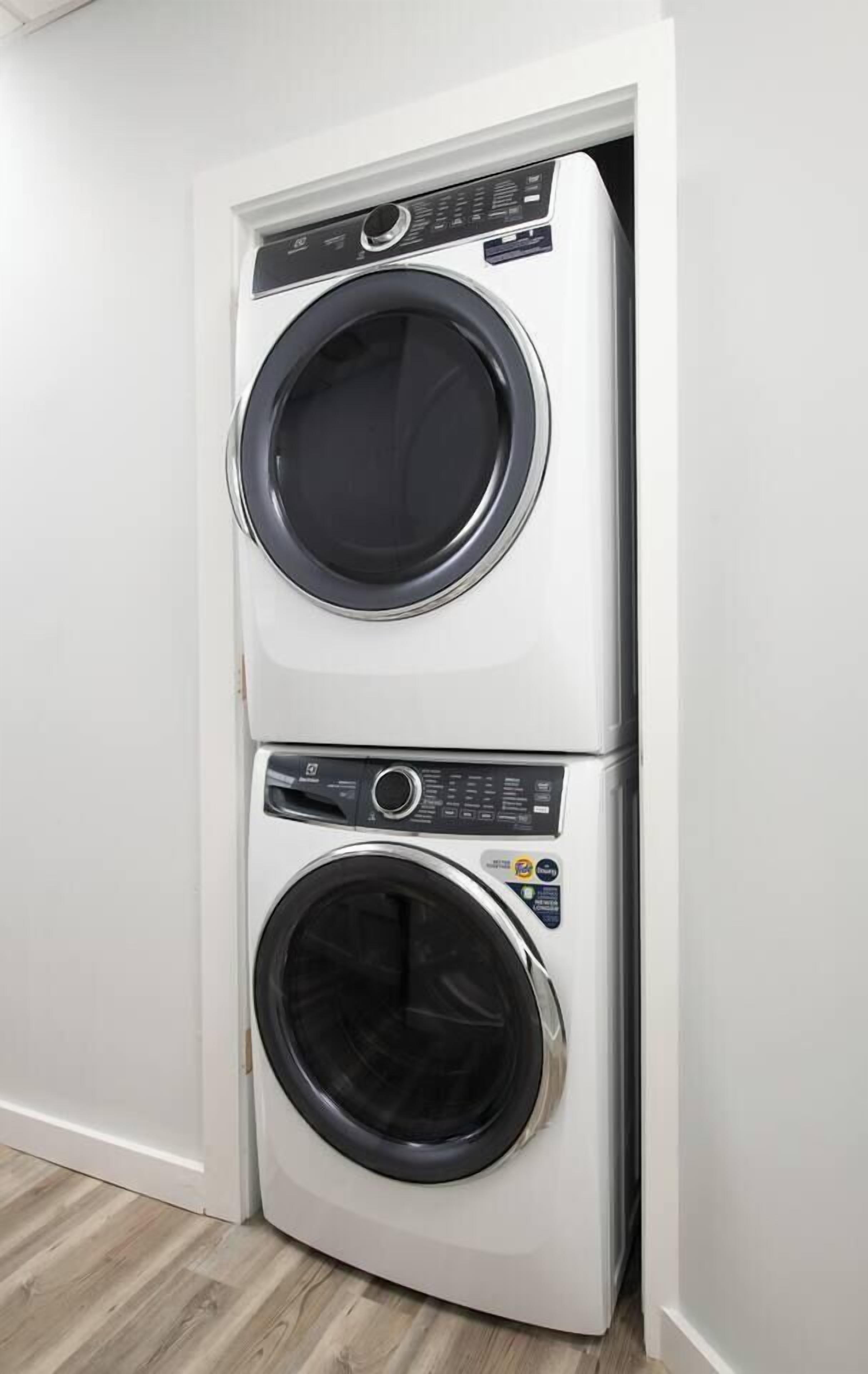 A stacked washer and dryer unit in a corner of a room with wooden flooring.