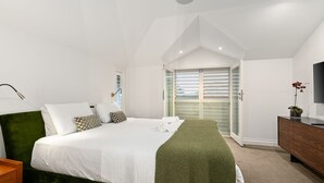 Ein modernes Schlafzimmer mit Bett, grünem Kopfteil, Nachttischlampe, Fernseher und einer Glaswand, die auf einen Balkon führt.