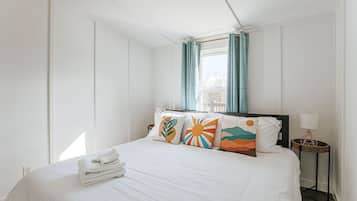 Un lit bien fait, avec du linge de lit blanc et des coussins colorés, une table de chevet avec une lampe et une fenêtre avec des rideaux.