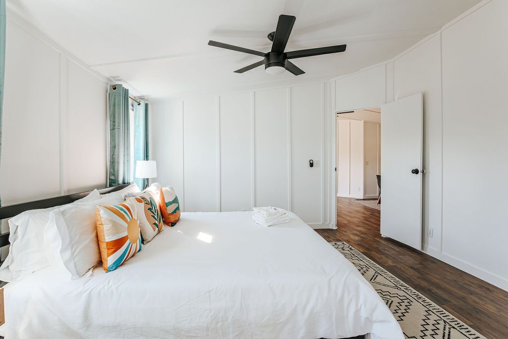 Une chambre à coucher comprenant un lit, des oreillers, un ventilateur de plafond, une fenêtre avec des rideaux et une porte donnant sur une autre pièce.