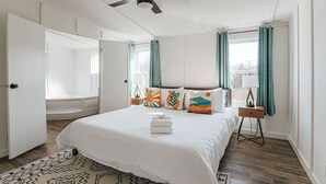 Une chambre avec un lit, un ventilateur de plafond, une fenêtre avec des rideaux, une table de chevet avec une lampe et une vue sur le paysage urbain.