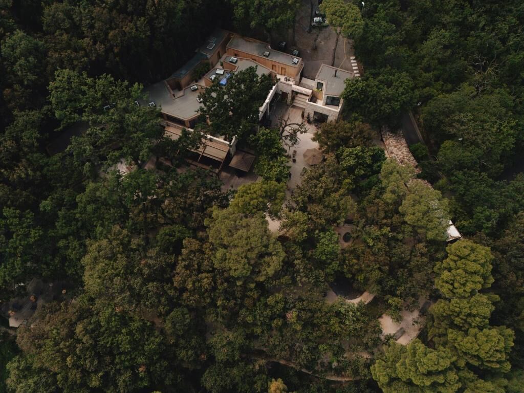 Una vista aerea di una struttura circondata da una fitta foresta, con un edificio centrale su più livelli, un'area patio e una piccola costruzione separata.