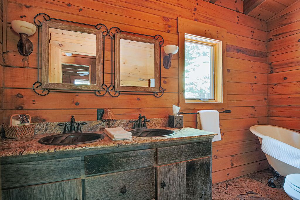 A rustic wooden cabin bathroom with a large vanity, two mirrors, a bathtub, and a window providing natural light.