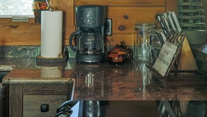 Het interieur van een houten hutje met een keukenhoek, inclusief een koffiezetapparaat, mokken en een glazen kast met wijnglazen en een klein decoratief object.