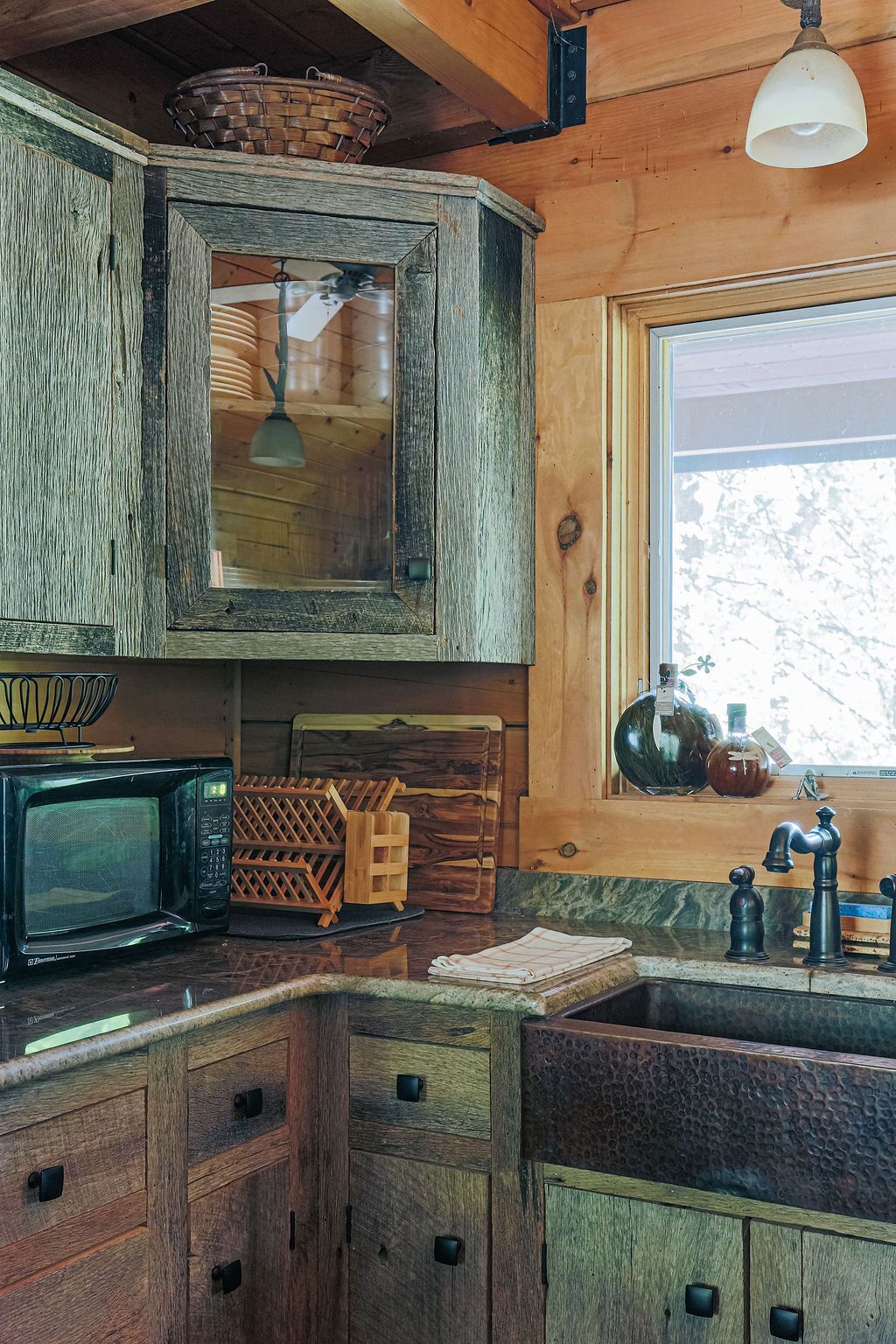 A rustic kitchen with wooden cabinets, a vintage microwave, and a window with a view of trees.