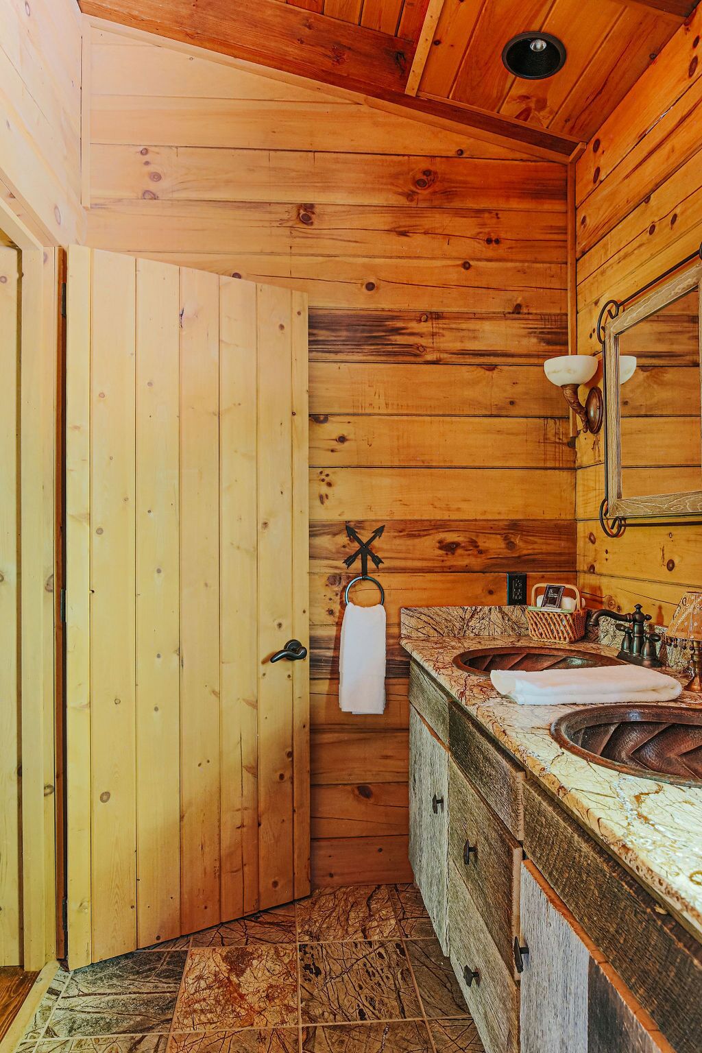 Un baño de cabaña de madera con una puerta de madera, una toalla colgando de un perchero, un lavabo con encimera de mármol y un espejo con un aplique.