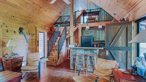 Een landelijk chaletinterieur met houten inrichting, een trap naar een vide, een keukenhoek en een bar met krukken.