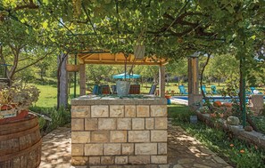 Eine Steinsitzbank unter einer Pergola mit einer hängenden Blumenampel und einem blauen Sonnenschirm.