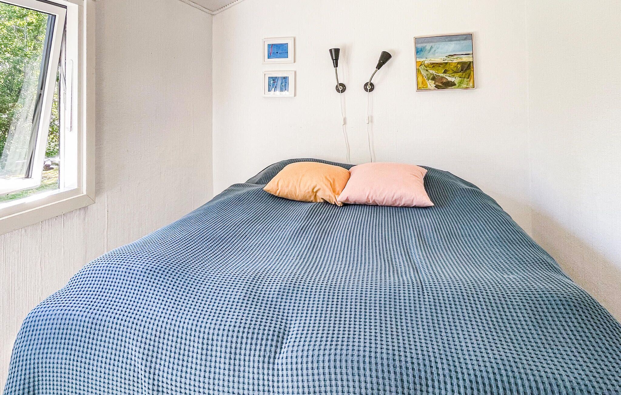 Un lit simple avec une couvre-lit à motifs bleus et deux oreillers, une fenêtre donnant sur la verdure, et deux tableaux encadrés accrochés au mur.