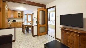 Een kamer met een eettafel, stoelen, een televisie, houten kasten en een keukenhoek.