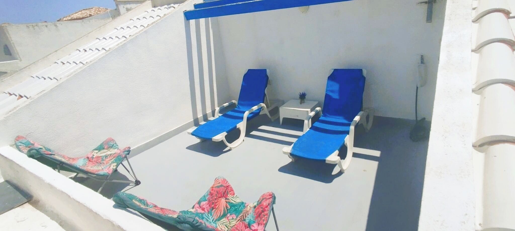 Une terrasse sur le toit, avec des fauteuils bleus et une petite table, entourée de murs blancs et d’une balustrade blanche.