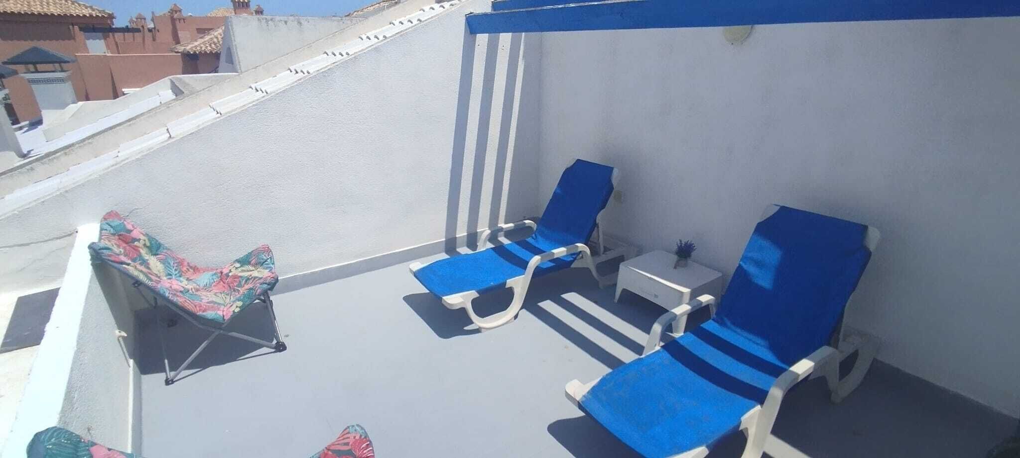 A rooftop terrace with blue lounge chairs, a small table, and a colorful chair.