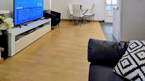 Un saló amb televisió, un sofà negre, taula de dinar amb cadires i paret blanca amb rellotge.