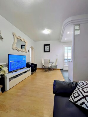 A living room with a TV, a black sofa, a dining table with chairs, and a white wall with a clock. -  APARTMENT NEXT TO THE BEACH A/C  ROOF TERRACE WIFI UK TV SPANISH (Torrevieja)