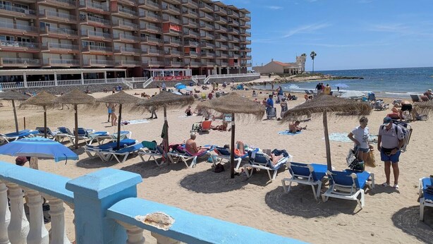 Un hotel a la vora de la platja amb nombroses balconades, una platja sorrenca amb gandules i ombrel·les, i una vista despejada de l'oceà.