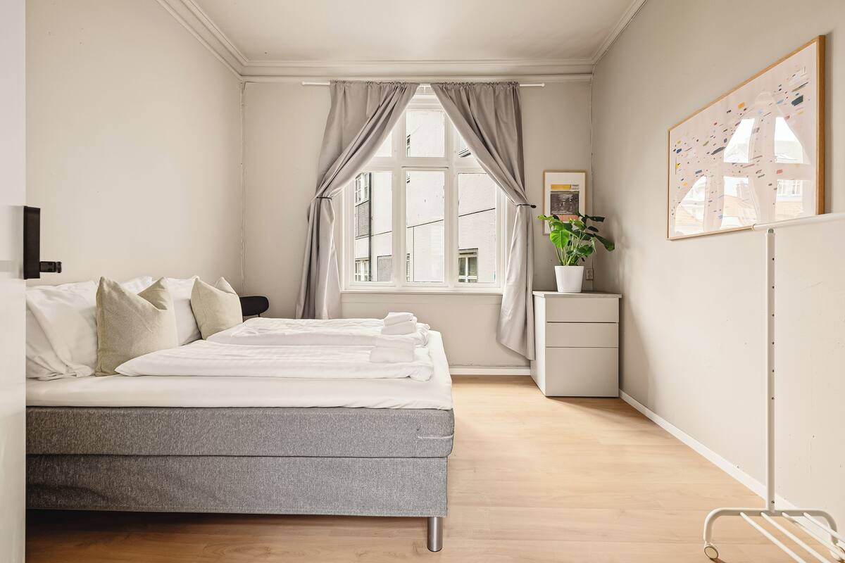 Un dormitorio con cama, una mesita de noche, una planta en maceta, una ventana con cortinas y una obra de arte colgada en la pared.