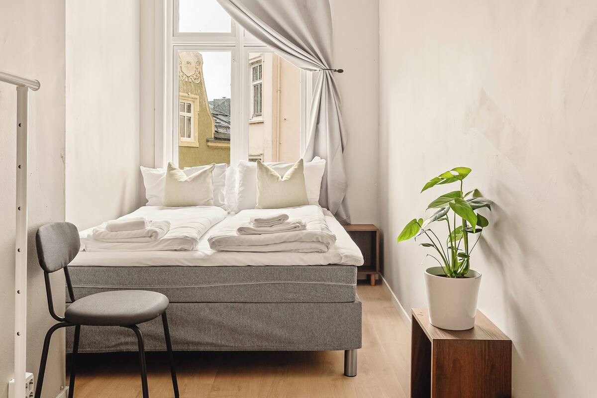 Un dormitorio con cama, una silla, una mesita de noche, una planta en maceta y una ventana con cortinas.