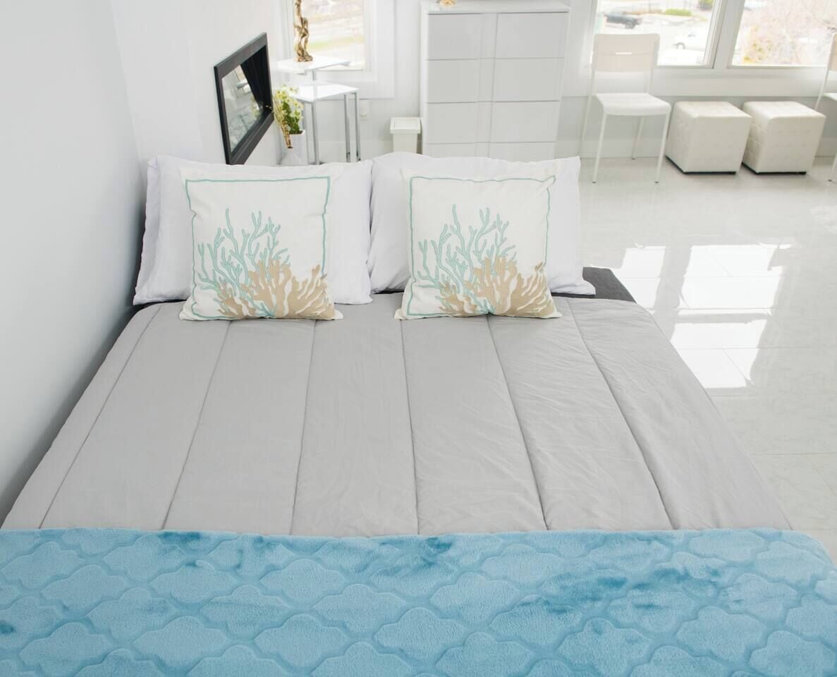 Una cama con colchón gris claro, dos almohadas estampadas en color coral y un corredor de cama azul con textura.