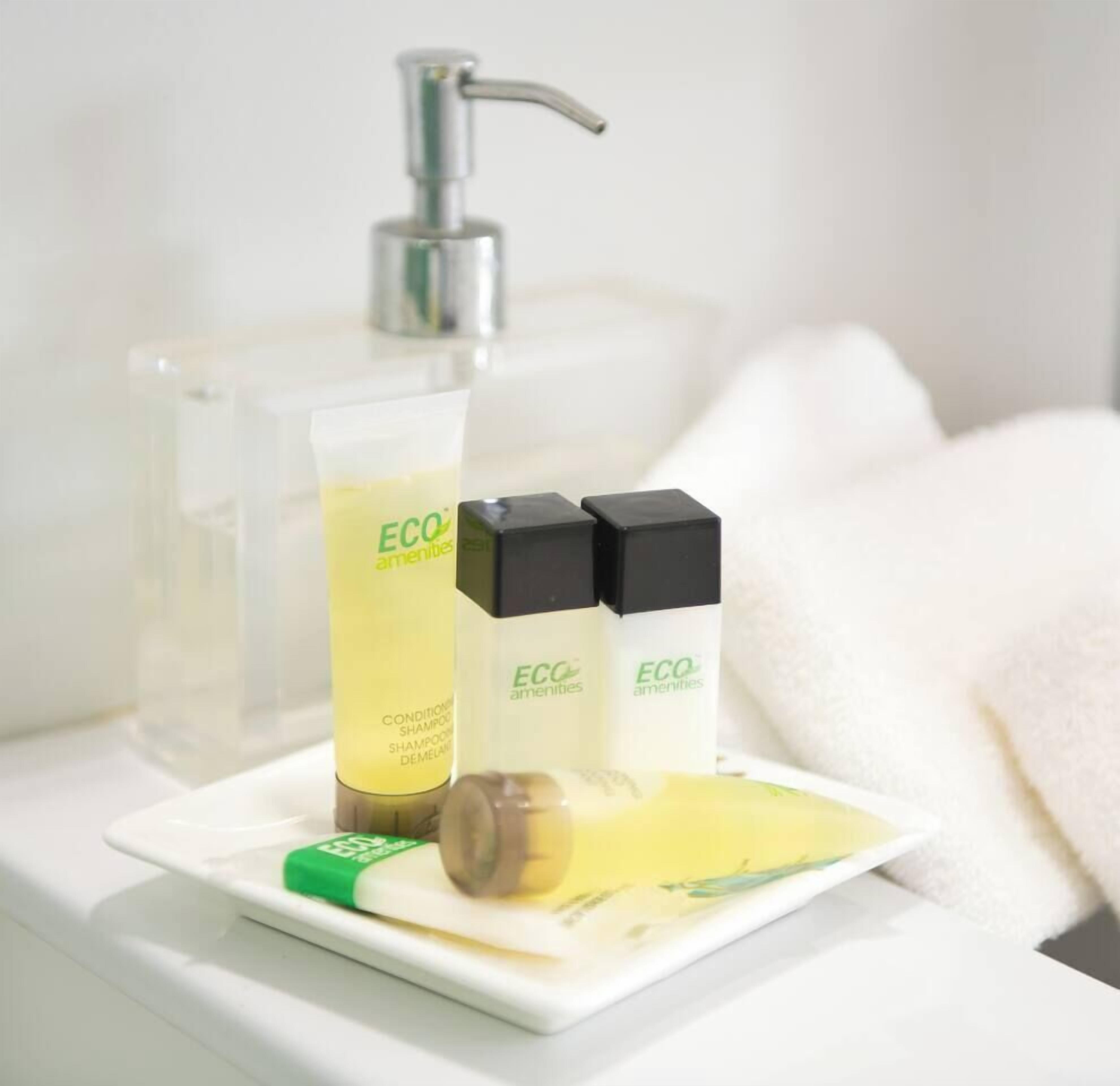 A bathroom with a clear glass bottle of shampoo, two black and white bottles of lotion, and a green bar of soap on a white tray.
