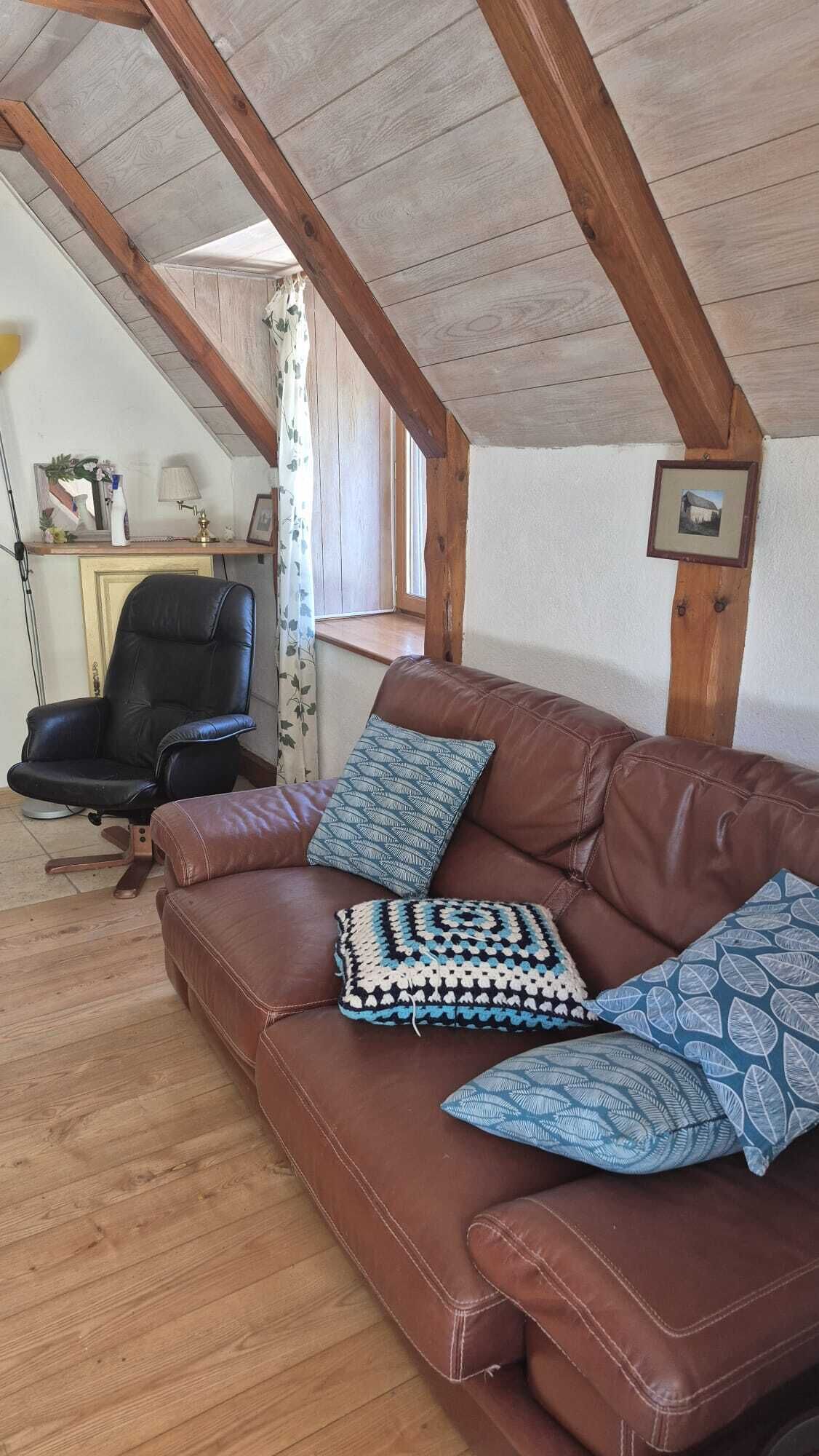 Ein gemütliches Zimmer mit einem braunen Ledersofa, einem schwarzen Bürostuhl und einer Holzbalkendecke.